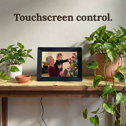 A 10-inch black Family Frame photo sharing frame resting on a natural wood table surrounded by cascading indoor plants. This premium WiFi photo frame provides an effortless user experience through a highly responsive, intuitive touchscreen control interface. Recognized as an ideal long-distance family gift, it offers unlimited users the ability to curate daily photo updates completely free of subscription charges.
