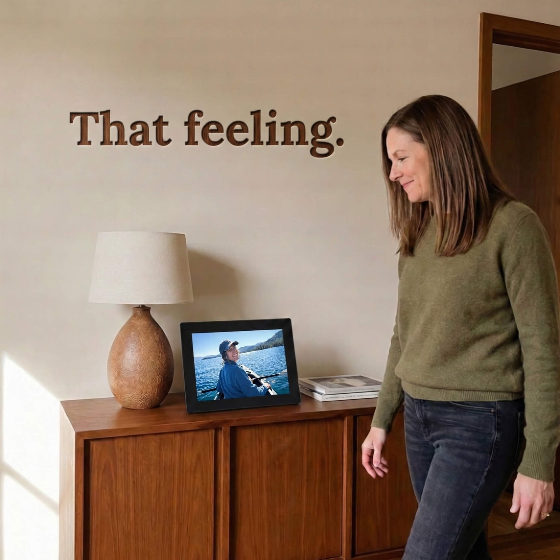 A 10-inch black Family Frame WiFi photo frame bringing a smile to a user standing beside a modern wooden cabinet. Engineered specifically as a heartwarming long-distance family gift, this sleek smart picture frame instantly receives new photos from loved ones living far away. It functions as a powerful digital memory display to consistently spark joy and combat loneliness through effortless photo sharing.
