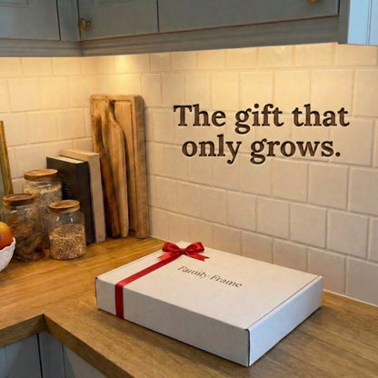 A 10-inch classic black Family Frame smart picture frame packaged elegantly in a white gift box with a red bow, sitting on a sunlit wooden kitchen counter. Perfectly presented as a luxury long-distance family gift, this beautifully boxed WiFi photo frame is built to foster daily emotional connections. The premium electronic photo frame serves as a continuous digital memory display, updating effortlessly through its dedicated secure application.
