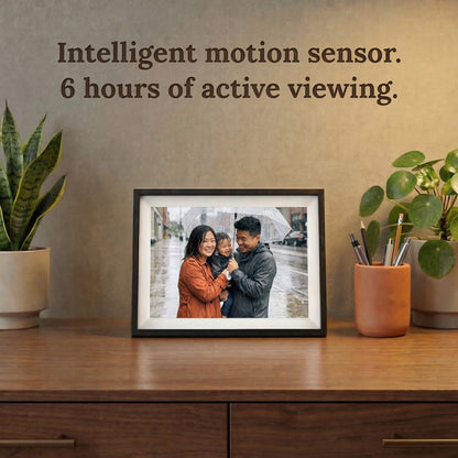 A 10-inch cordless classic black Family Frame smart picture frame with a classic white mat resting on a dark wood credenza surrounded by houseplants. Highlighting its completely cordless design, this electronic photo frame features an intelligent motion sensor and rechargeable battery for up to 6 hours of active, wire-free viewing. This innovative WiFi photo frame is the perfect long-distance family gift, allowing memories to be displayed on any surface without worrying about nearby outlets.
