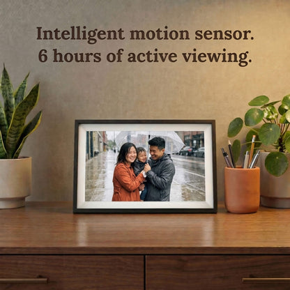 A 10-inch cordless classic black Family Frame smart picture frame with a classic white mat resting on a dark wood credenza surrounded by houseplants. Highlighting its completely cordless design, this electronic photo frame features an intelligent motion sensor and rechargeable battery for up to 6 hours of active, wire-free viewing. This innovative WiFi photo frame is the perfect long-distance family gift, allowing memories to be displayed on any surface without worrying about nearby outlets.