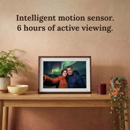 A 10-inch cordless mocha wood Family Frame smart picture frame with a classic white mat resting on a wooden entryway table surrounded by indoor plants. Highlighting its completely cordless design, this electronic photo frame features an intelligent motion sensor and rechargeable battery for up to 6 hours of active, wire-free viewing. This innovative WiFi photo frame is the perfect long-distance family gift, allowing memories to be displayed on any surface without worrying about nearby outlets.
