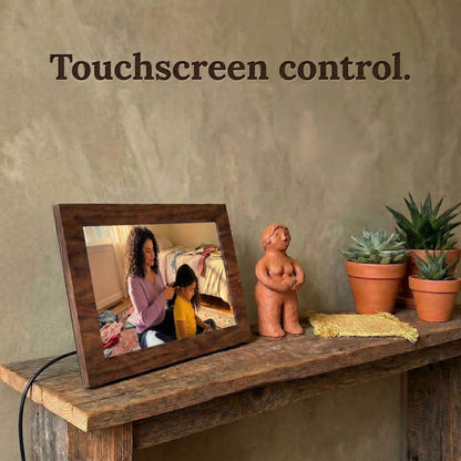 A 10-inch oak wood Family Frame photo sharing frame resting on a weathered rustic table surrounded by small succulent plants. This high-end WiFi photo frame provides an effortless user experience through a highly responsive, intuitive touchscreen control interface. Recognized as an ideal long-distance family gift, it offers unlimited users the ability to curate daily photo updates completely free of subscription charges.