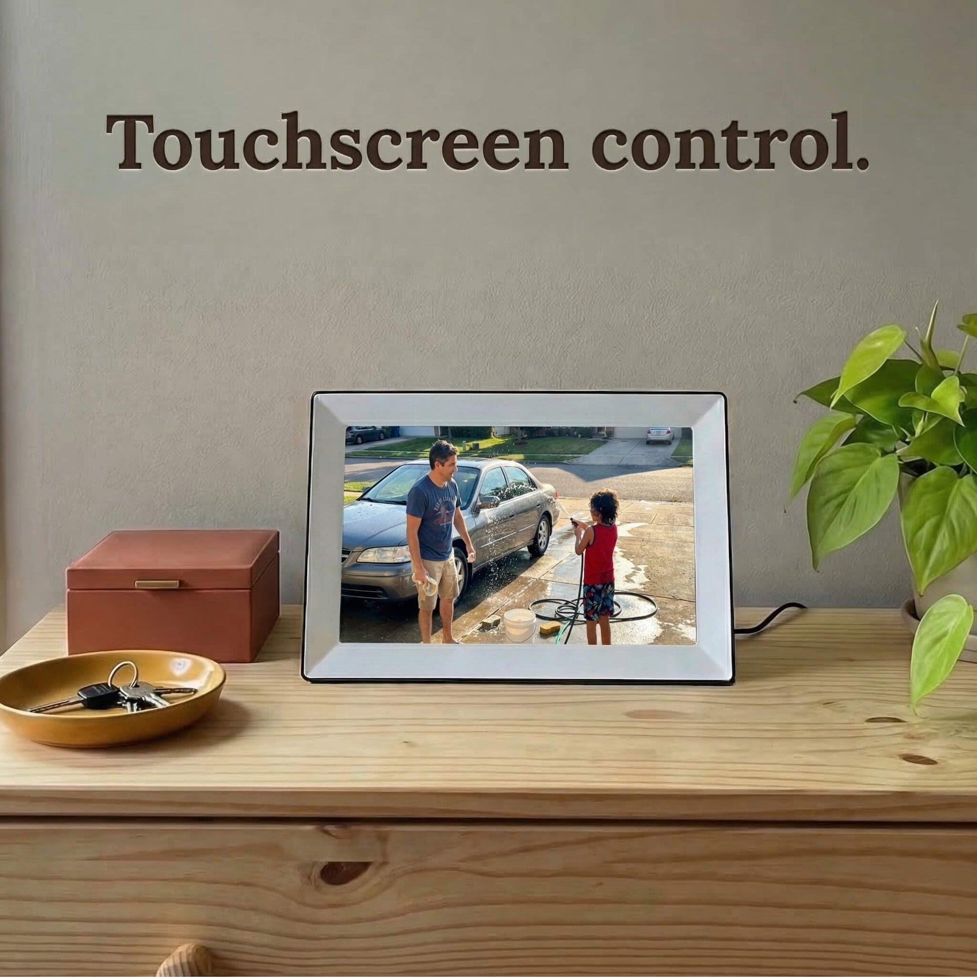 A 10-inch white Family Frame photo sharing frame resting on a natural wood surface next to modern home decor. This premium WiFi photo frame includes responsive touchscreen control, making it effortless to swipe through galleries, adjust settings, and favorite cherished family moments. Serving as a top-rated long-distance family gift, the digital display requires zero subscription fees to operate and enjoy.
