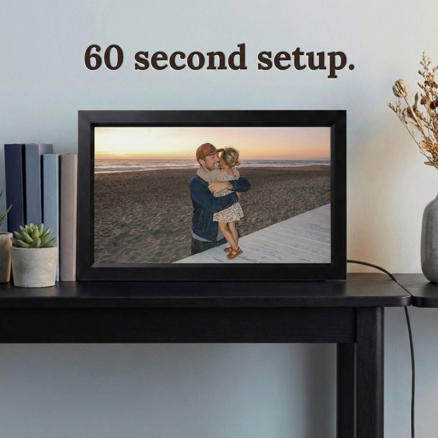 A 16-inch black Family Frame electronic photo frame displayed prominently on a dark wooden console table beside decorative books. Prioritizing accessibility, this intuitive smart picture frame boasts a frictionless, easy 60-second WiFi setup process that anyone can master in minutes. This completely no subscription fee digital frame ensures immediate and uninterrupted photo sharing for families of all technical backgrounds.
