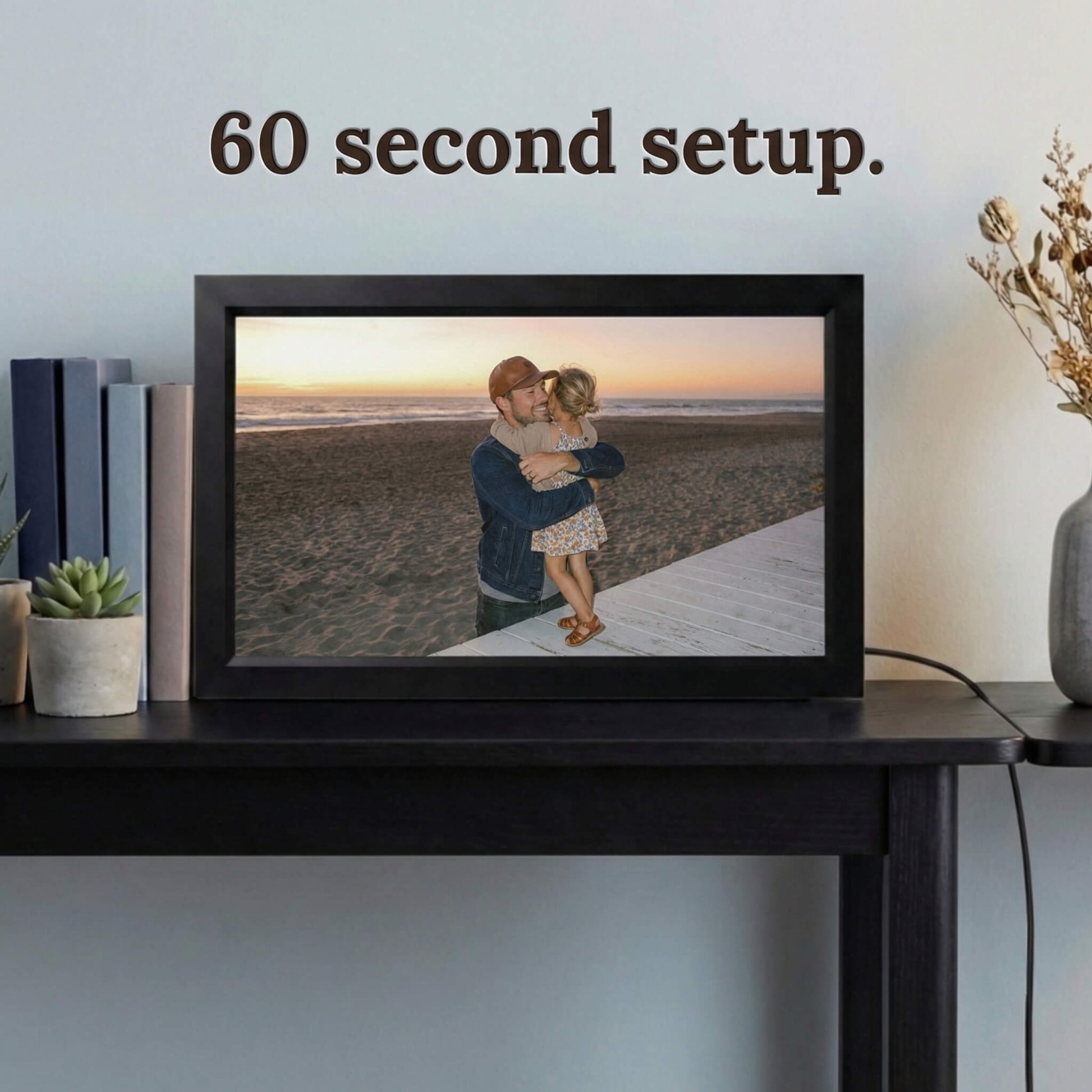 A 16-inch black Family Frame electronic photo frame displayed prominently on a dark wooden console table beside decorative books. Prioritizing accessibility, this intuitive smart picture frame boasts a frictionless, easy 60-second WiFi setup process that anyone can master in minutes. This completely no subscription fee digital frame ensures immediate and uninterrupted photo sharing for families of all technical backgrounds.
