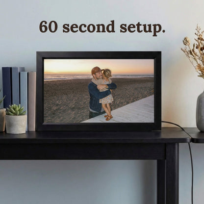A 16-inch black Family Frame electronic photo frame displayed prominently on a dark wooden console table beside decorative books. Prioritizing accessibility, this intuitive smart picture frame boasts a frictionless, easy 60-second WiFi setup process that anyone can master in minutes. This completely no subscription fee digital frame ensures immediate and uninterrupted photo sharing for families of all technical backgrounds.
