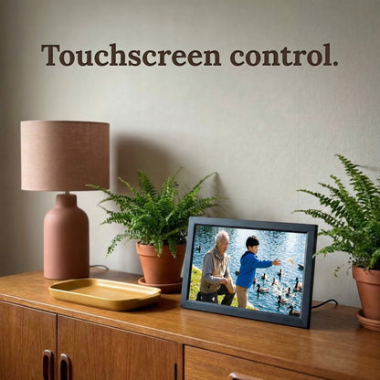A 16-inch black Family Frame photo sharing frame resting on a mid-century modern credenza near a textured lamp. This premium WiFi photo frame provides an effortless user experience through a highly responsive, intuitive touchscreen control interface. Recognized as an ideal long-distance family gift, it offers unlimited users the ability to curate daily photo updates completely free of subscription charges.
