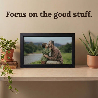 A 16-inch black Family Frame smart picture frame situated on a smooth beige shelf alongside modern potted plants. Designed to elevate home decor effortlessly, this electronic photo frame delivers an unparalleled high-resolution display that beautifully highlights the vibrant details of every uploaded image. Acting as a dedicated digital memory display, it bridges physical distance between family members with a constant stream of new photos.
