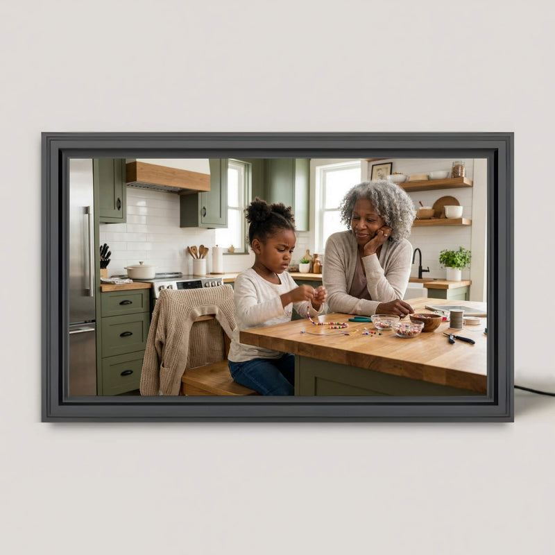 16 inch grey smart picture frame resting on a white surface. The screen displays a heartwarming photo of a grandmother and granddaughter making beaded jewelry together at a green kitchen island.
