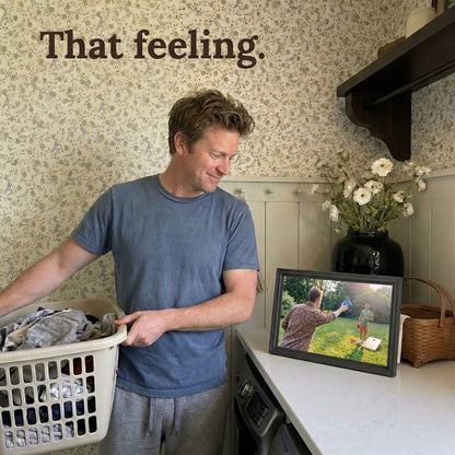 A 16-inch studio black Family Frame WiFi photo frame placed on a kitchen counter, bringing a smile to a user's face during everyday routines. Engineered as an incredibly thoughtful long-distance family gift, this easy setup smart frame instantly receives new photos from anywhere in the world. The digital memory display acts as a continuous source of joy, securely connecting families in real-time.
